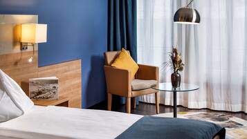 Una habitación de hotel con una cama, una mesita de noche de madera con una lámpara, una silla con un cojín amarillo, una pequeña mesa con un florero y un amplio ventanal con cortinas.
