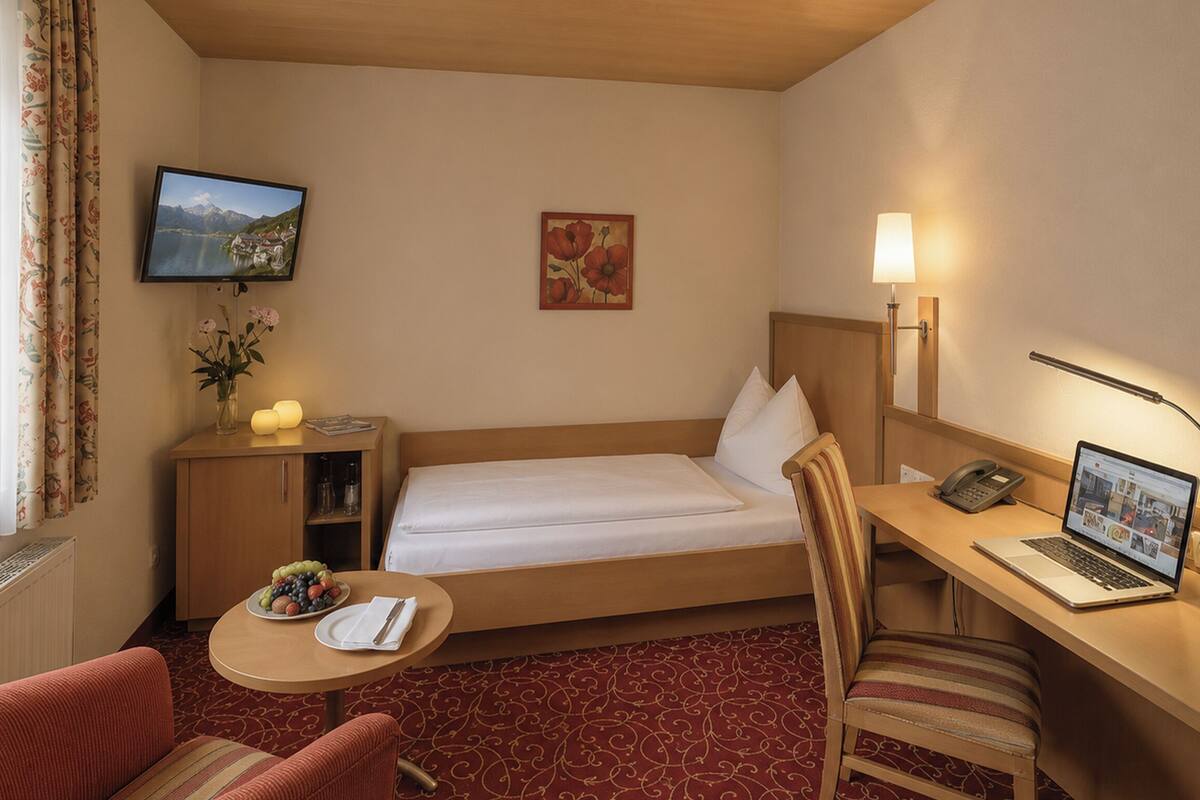 Una habitación de hotel con una cama, un escritorio con una computadora portátil, una silla, una mesita con frutero, un televisor y un cuadro en la pared.