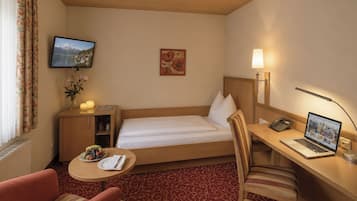 Ein Hotelzimmer mit einem Bett, einem Schreibtisch mit Laptop, einem Sessel, einem kleinen Tisch mit Obstschale, einem Fernseher und einem Bild an der Wand.