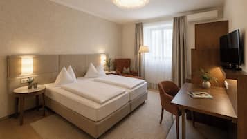 A hotel room with a double bed, a desk with a chair, a television, and a chandelier.