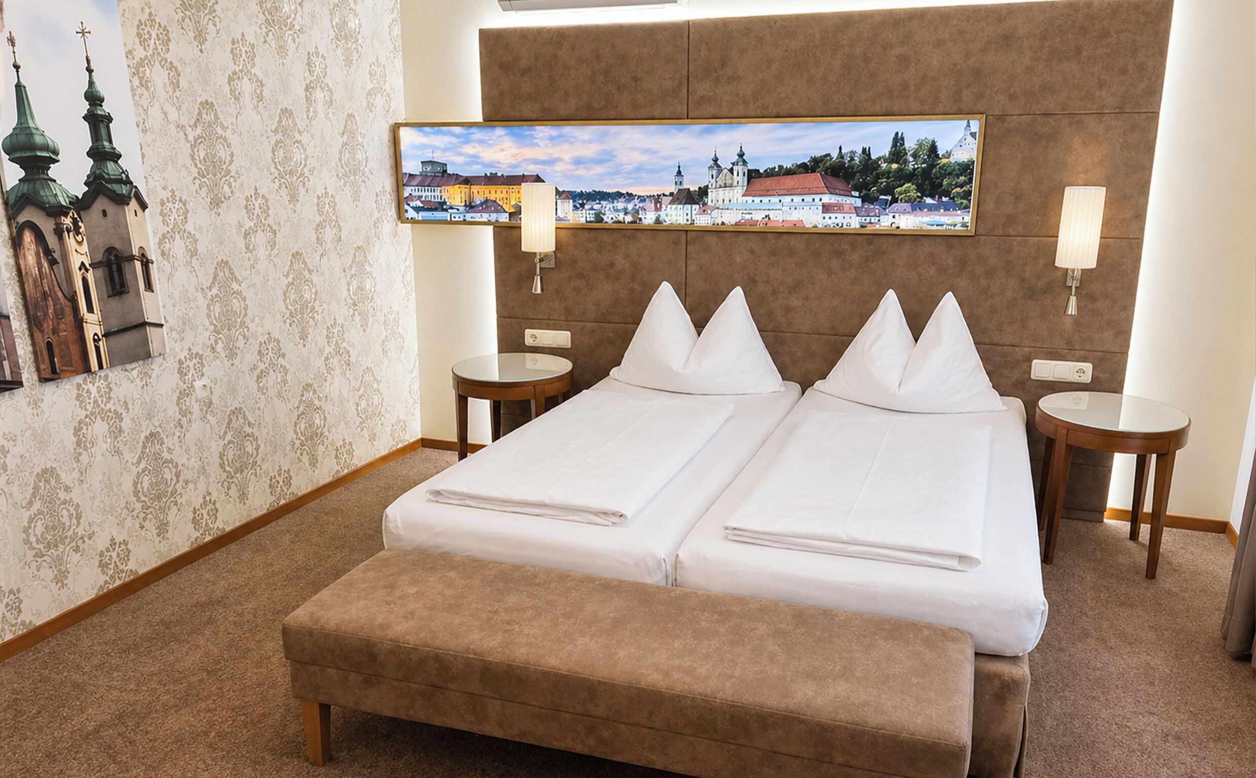 Una cama doble con dos almohadas, una mesita de noche con lámpara, un cuadro de un paisaje urbano y un papel tapiz estampado.