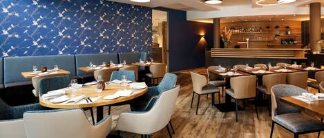 A modern restaurant with round tables, blue upholstered booths, and a bar area with a wooden counter.