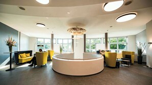 Una moderna zona de recepción con un mostrador circular central, asientos amarillos, ventanas amplias e iluminación empotrada en el techo.