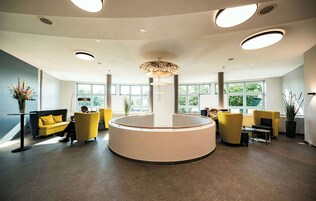 A modern reception area with a central circular desk, yellow seating, large windows, and a ceiling with recessed lighting.