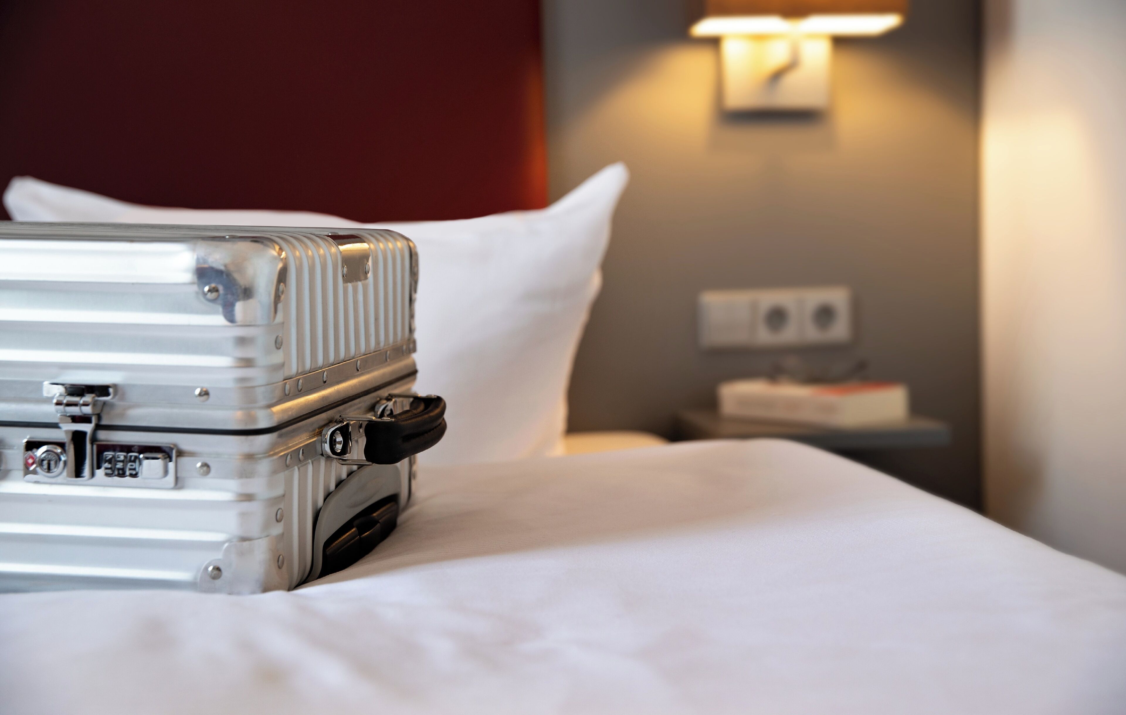 A neatly made bed with a suitcase on it, a white pillow, a bedside table with a lamp, and a wall socket.