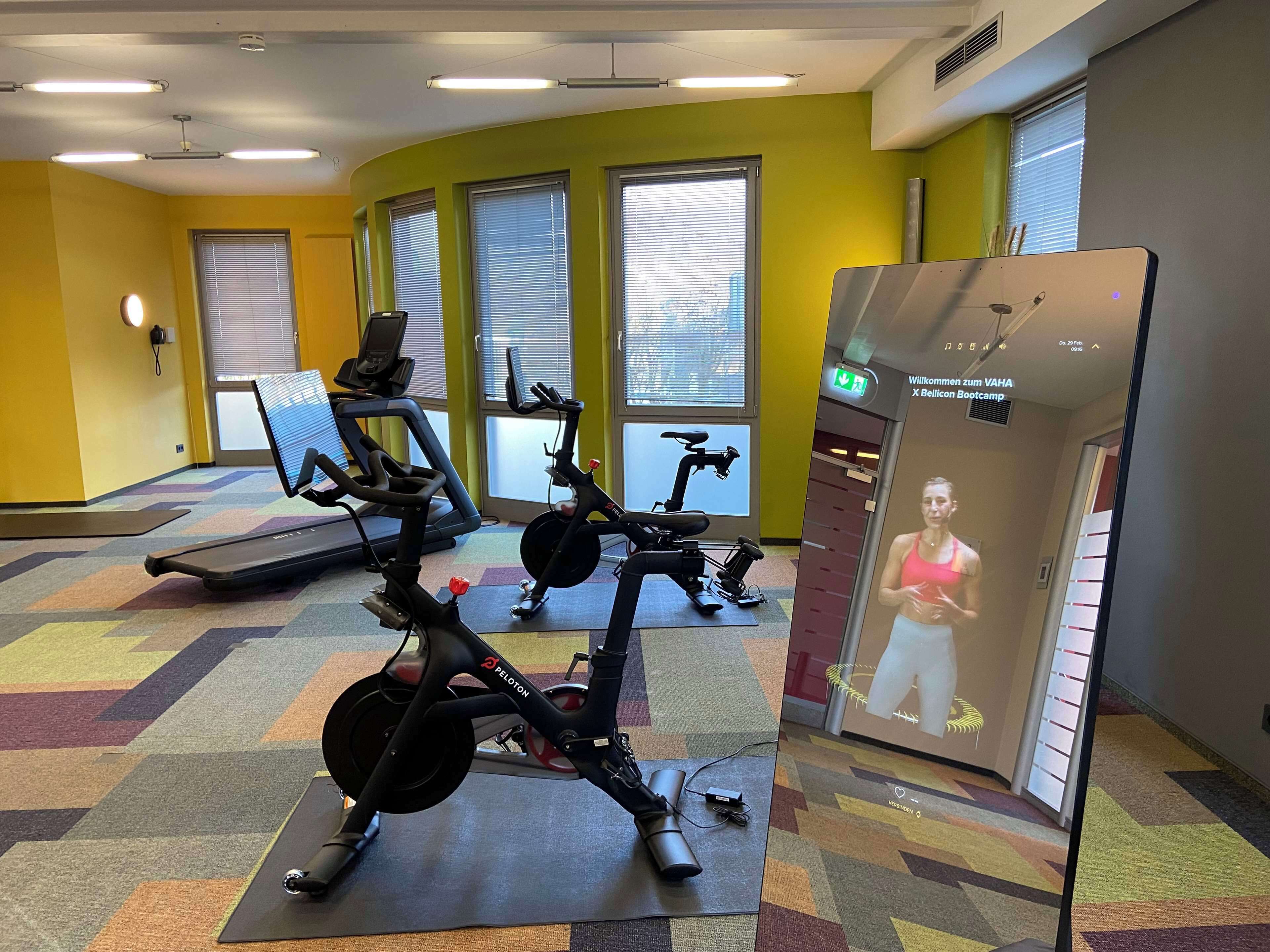 A gym with exercise equipment, a treadmill, and a stationary bike. There are windows with blinds, a wall-mounted phone, and a digital screen displaying a person.