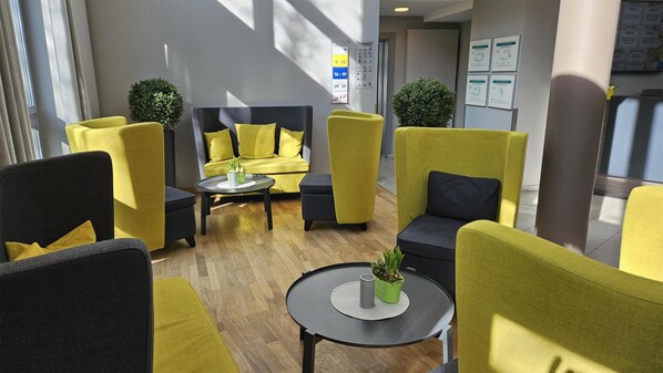 Un área de descanso moderna con mobiliario amarillo y negro, una mesa redonda con una planta y un ventanal que proporciona luz natural.