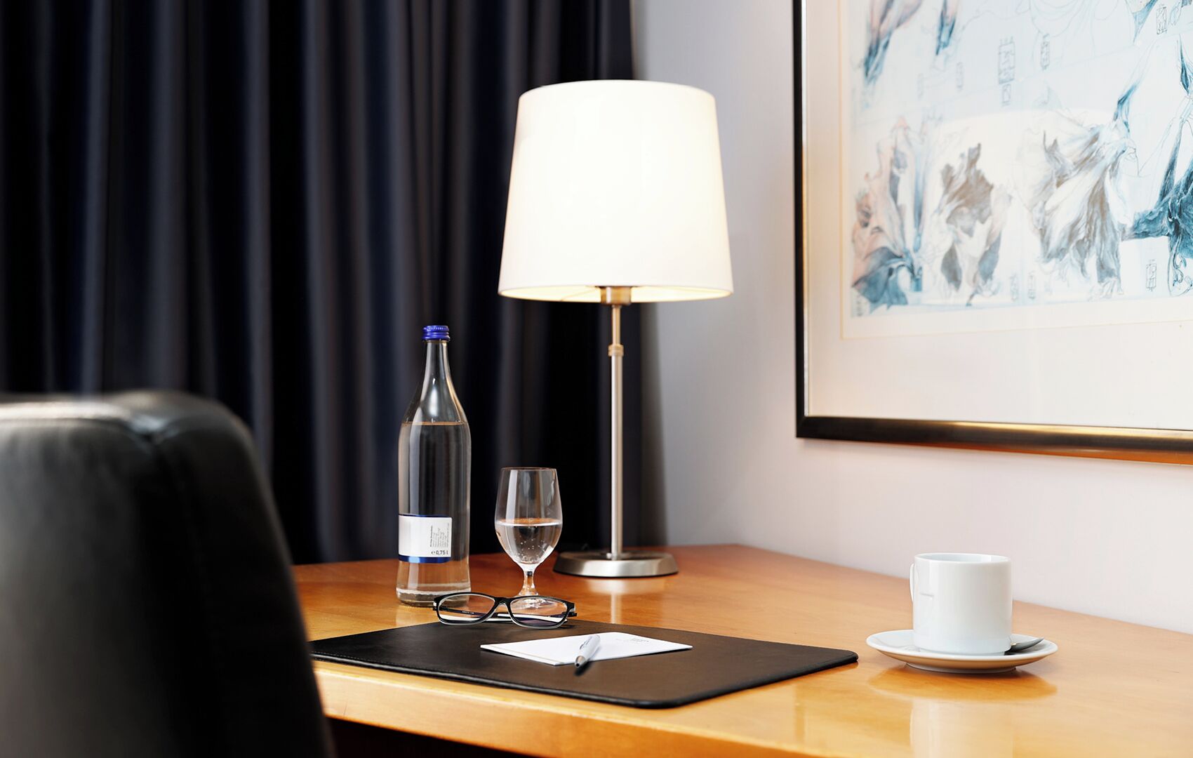 Une chambre d’hôtel avec un bureau en bois, un fauteuil en cuir noir, une lampe de table blanche, une bouteille de vin, un verre à vin, une œuvre d’art encadrée et une tasse blanche posée sur une soucoupe.