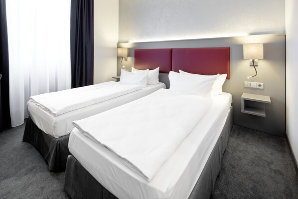 Habitación de hotel con dos camas individuales, cabecera roja, lámparas de noche y una pequeña estantería.