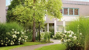 Un sendero conduce a un edificio de estilo arquitectónico moderno, rodeado de exuberante follaje y plantas con flores blancas.