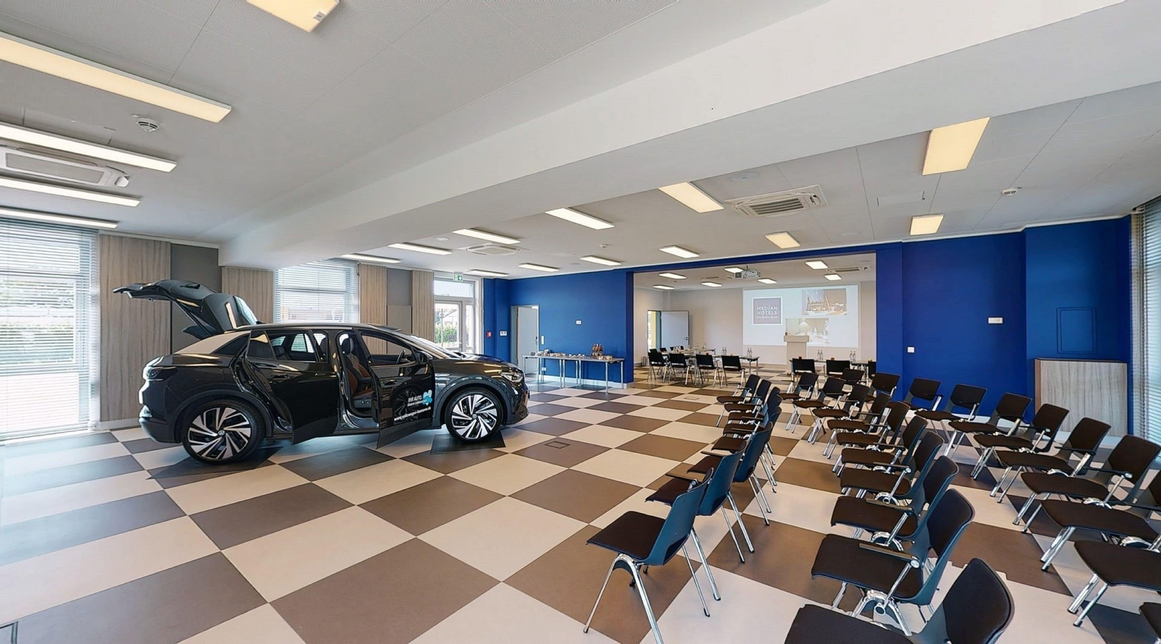 Salón de conferencias de hotel con un automóvil negro estacionado en primer plano, piso a cuadros, filas de sillas y una pantalla de presentación.