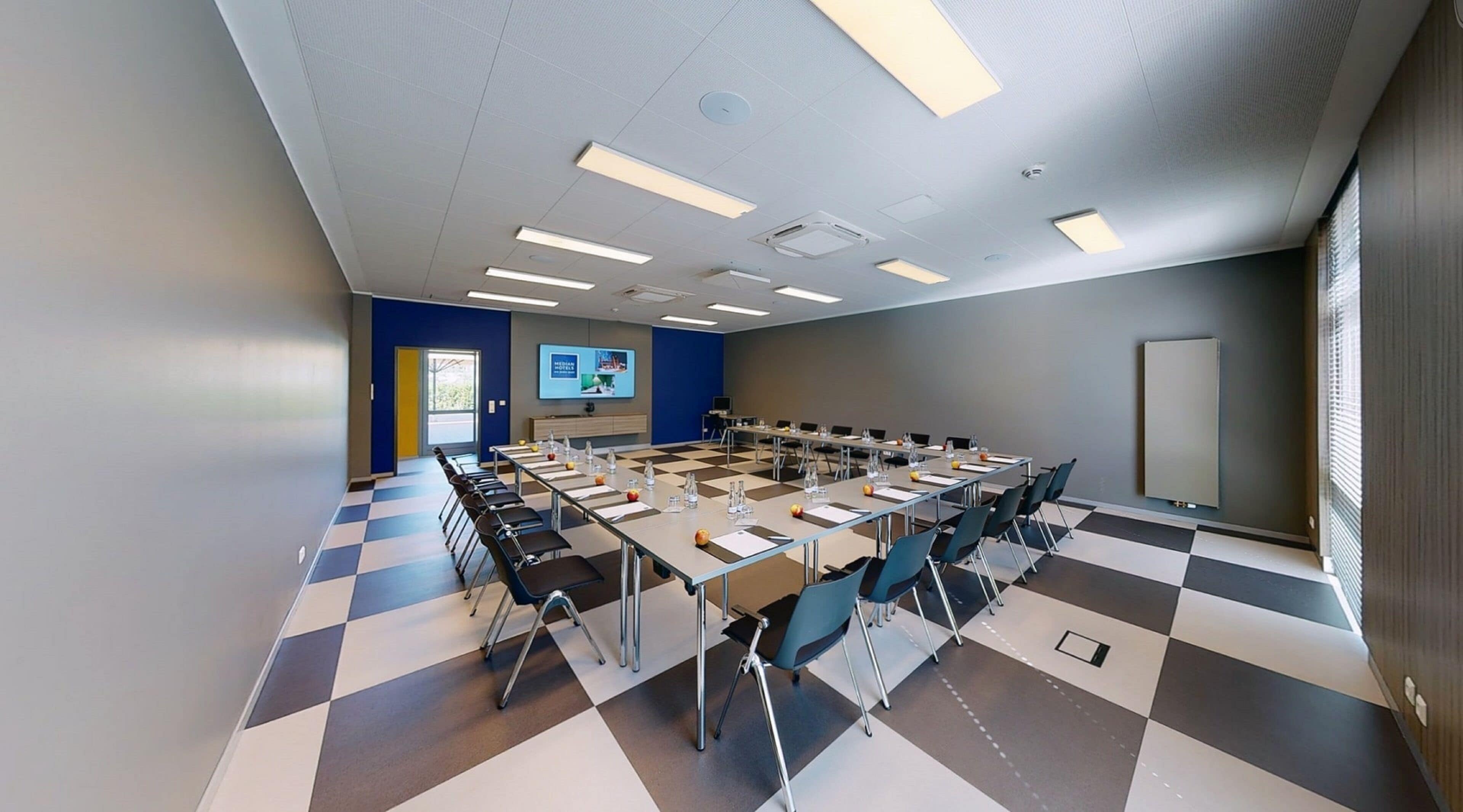 Sala de conferências com piso xadrez, várias mesas, cadeiras e um projetor.