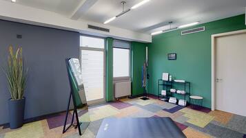 Una habitación con una alfombra colorida, una esterilla de yoga, un escritorio de pie, una planta en maceta y un estante con toallas.