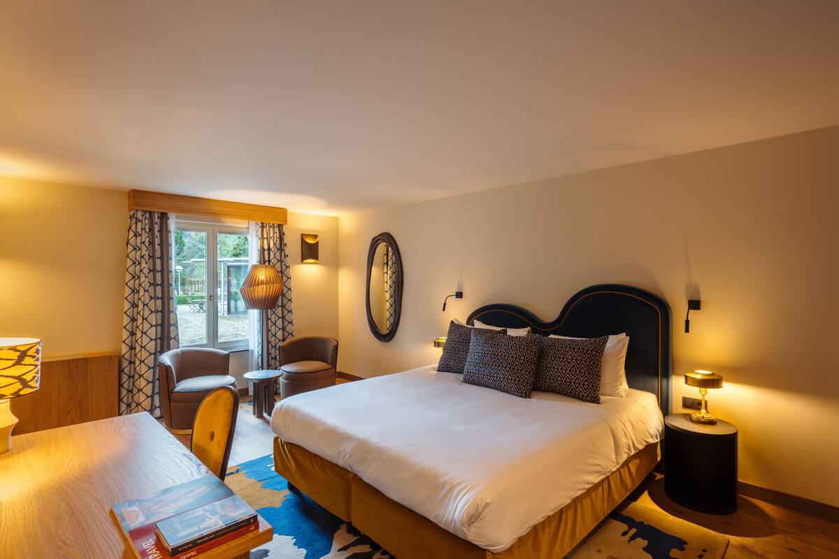Une chambre d’hôtel avec un lit, des tables de chevet, une chaise, un bureau et une fenêtre avec des rideaux.