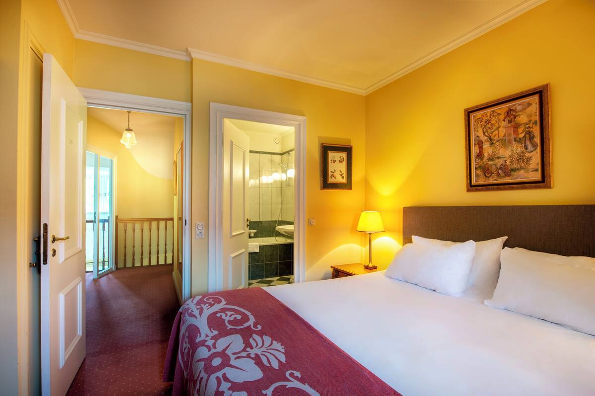 Une chambre d’hôtel comprenant un lit, une table de chevet, une lampe, un tableau encadré et une porte donnant accès à une salle de bain.