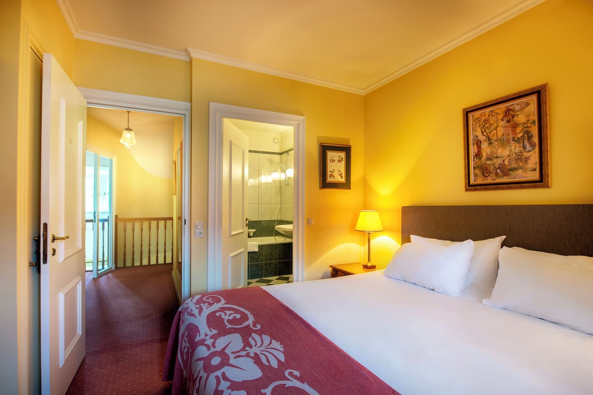 A hotel room with a bed, a nightstand, a lamp, a framed picture, and a door leading to a bathroom.