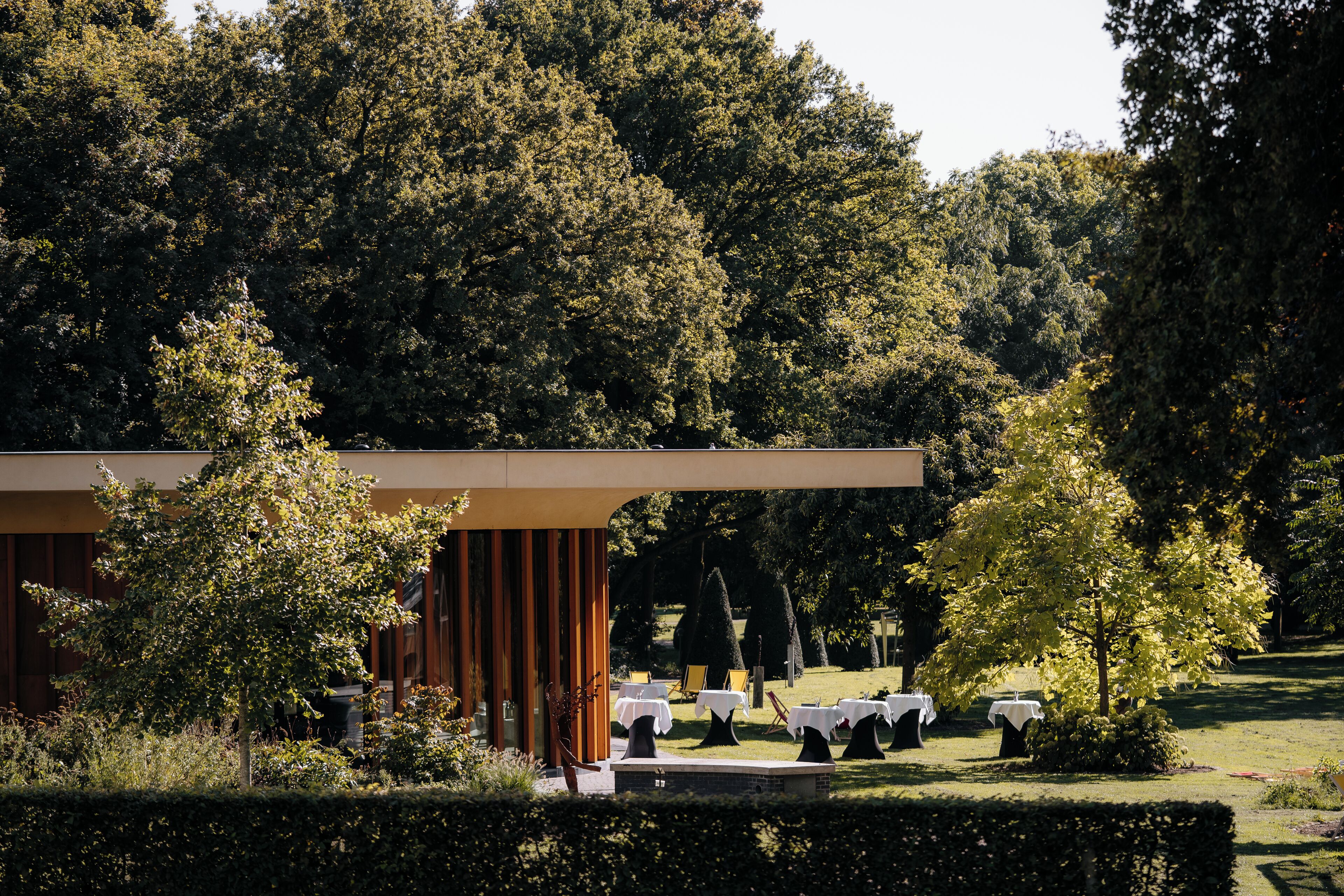 Een modern gebouw met een overdekt terras, omringd door bomen en verzorgd gazon.