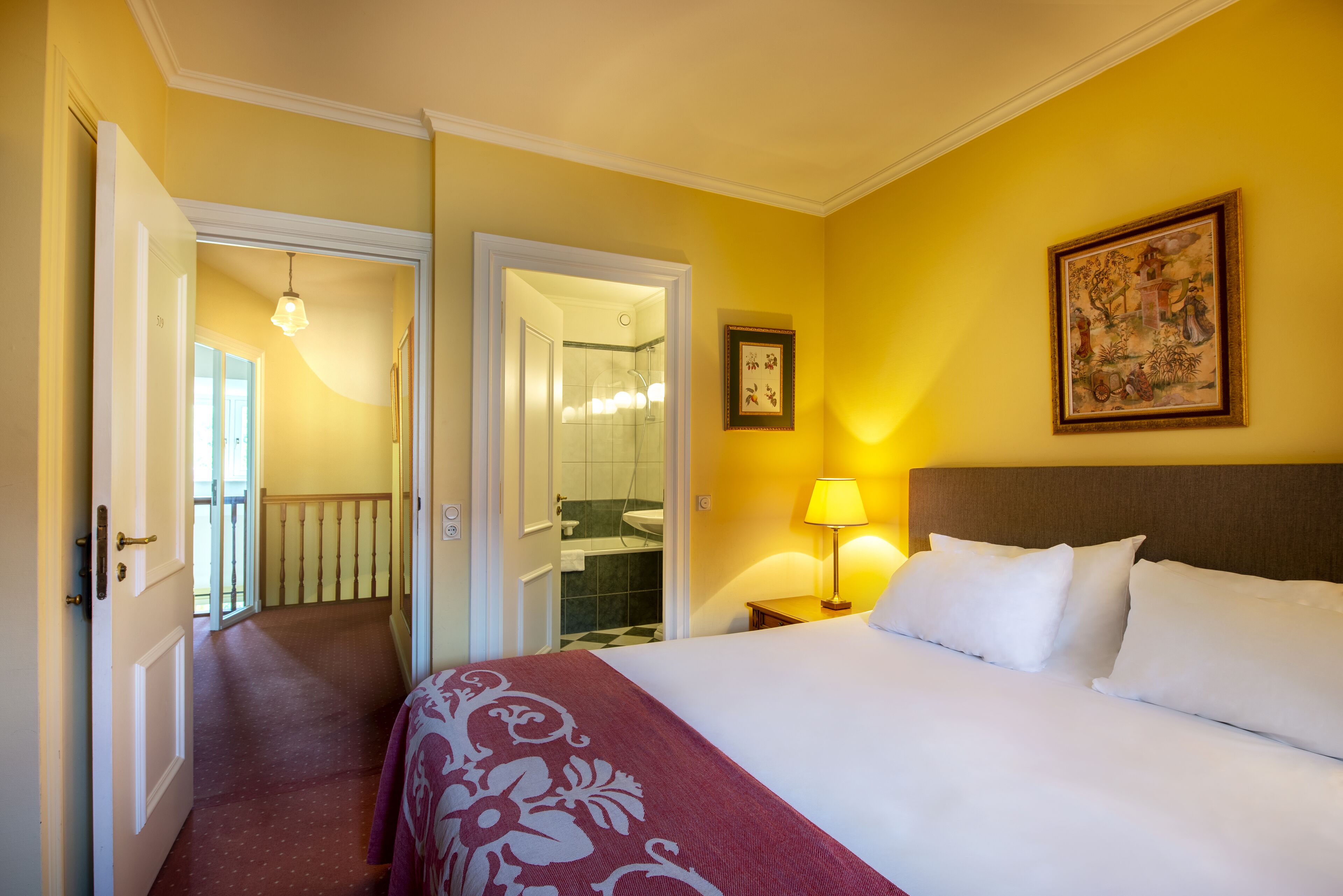 Una camera d'albergo con un letto, un comodino, una lampada, un quadro incorniciato e una porta che dà su un bagno.