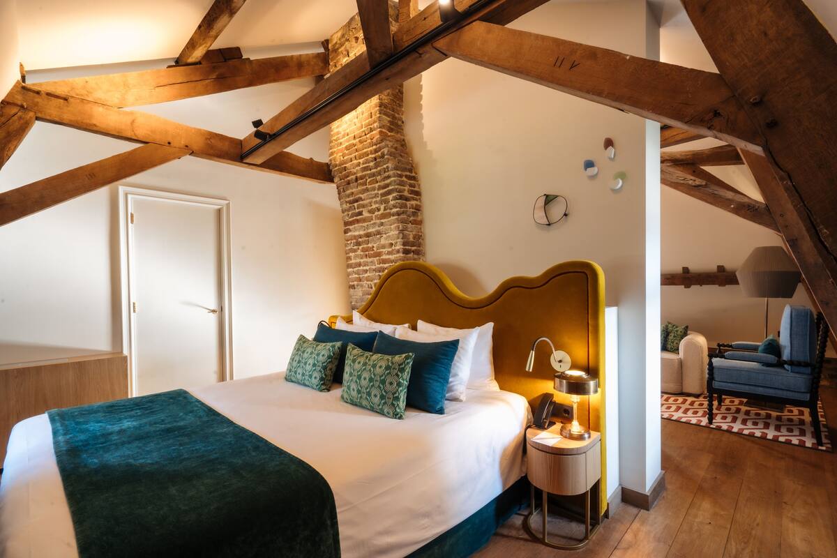 Une chambre avec un grand lit, un mur en briques, des poutres en bois, une table de chevet avec une lampe et une chaise.
