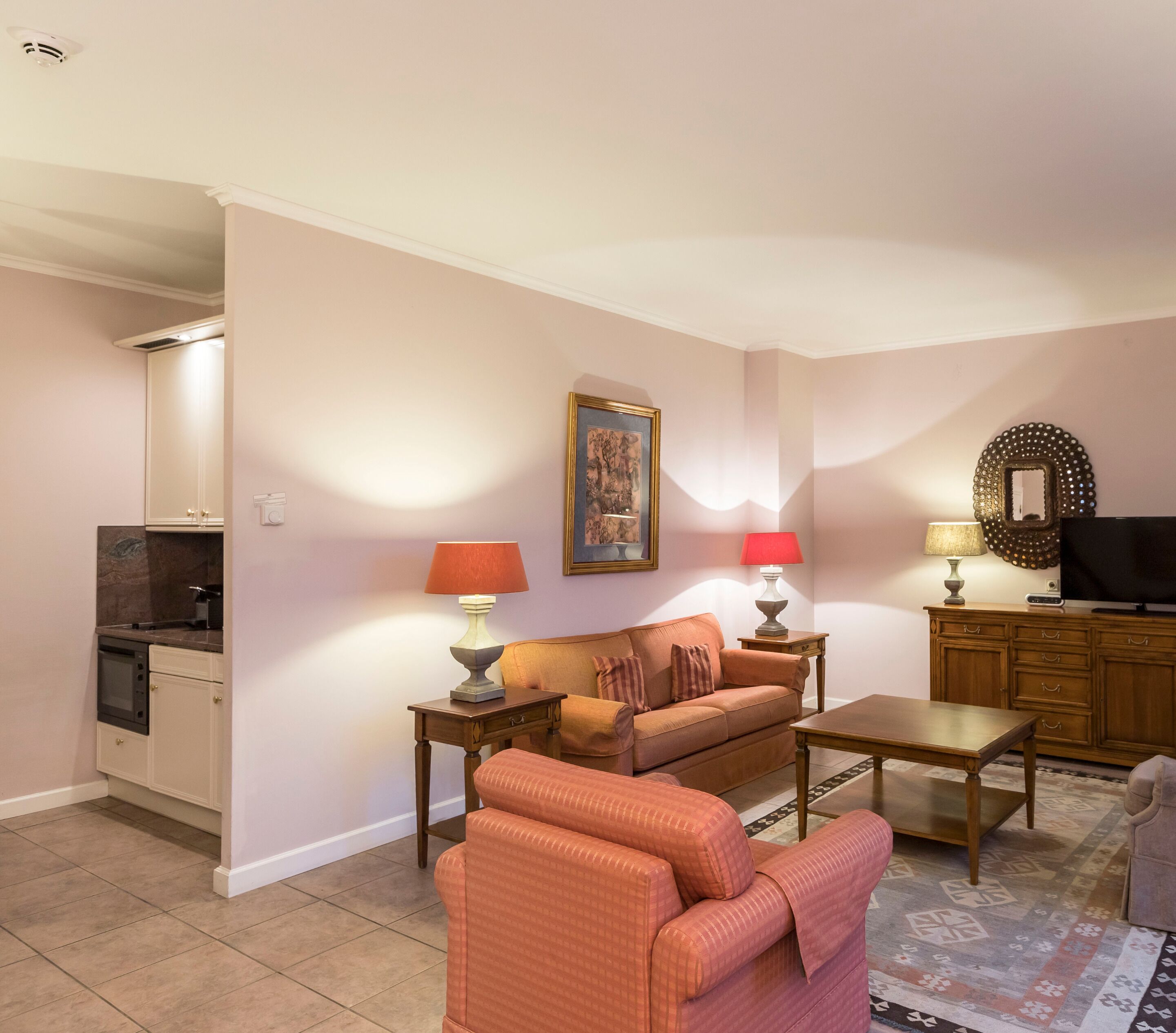 A living room with a sofa, two side tables with lamps, a television, and a kitchen area with cabinets and a microwave.