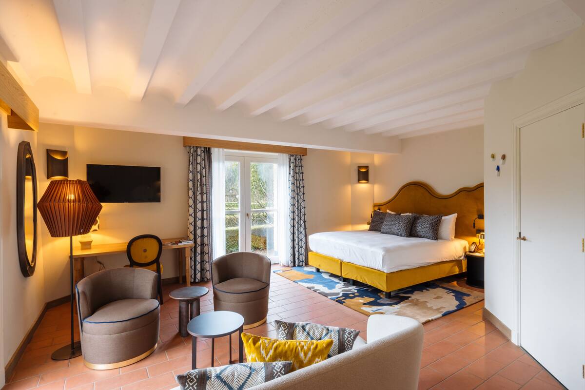 Une chambre d’hôtel avec un lit, deux fauteuils, une petite table et un canapé. Il y a une fenêtre avec des rideaux, un téléviseur à écran plat fixé au mur et une lampe sur une table d’appoint.