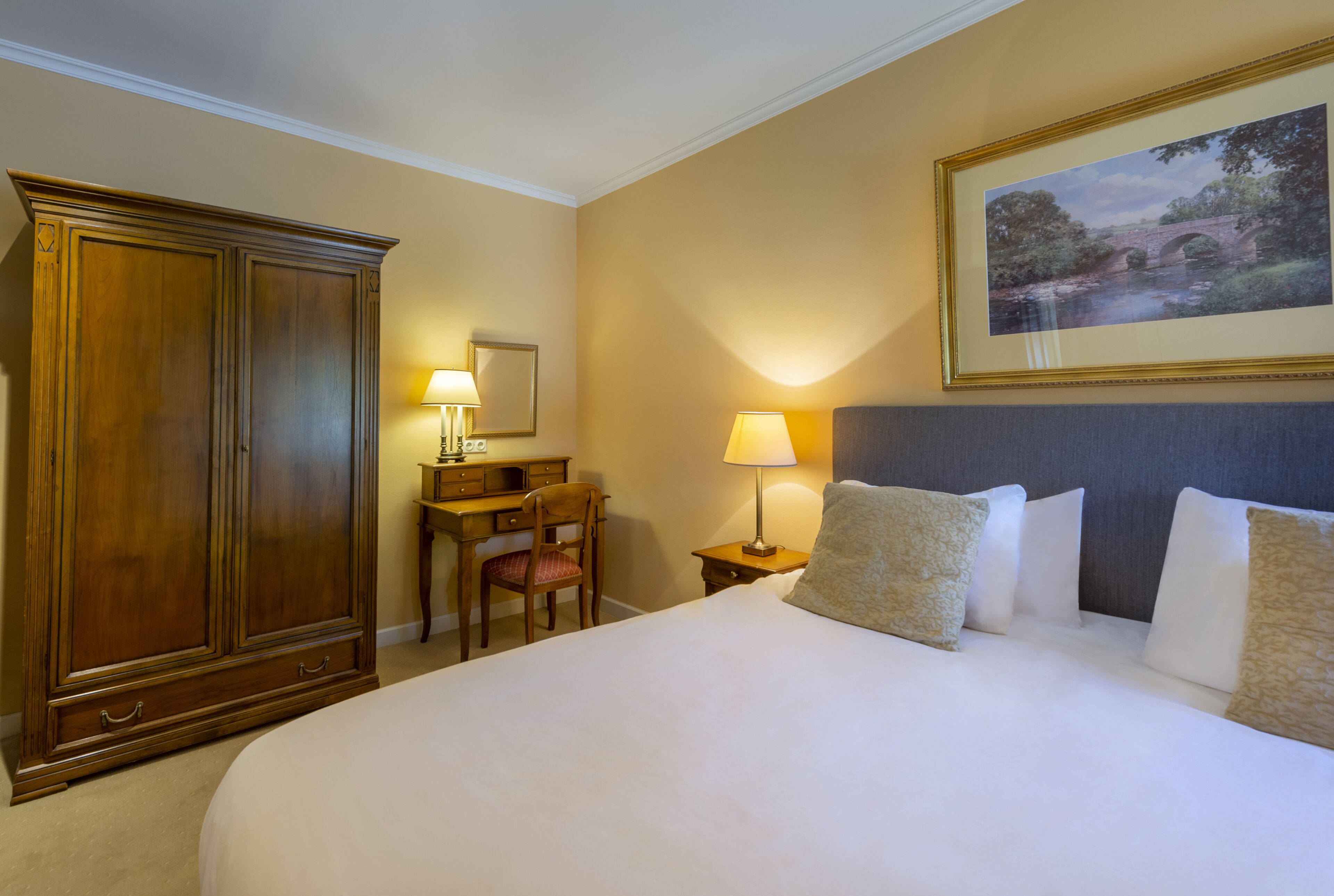 Habitación de hotel con una cama grande, un armario de madera, un escritorio con una silla y un cuadro enmarcado en la pared.