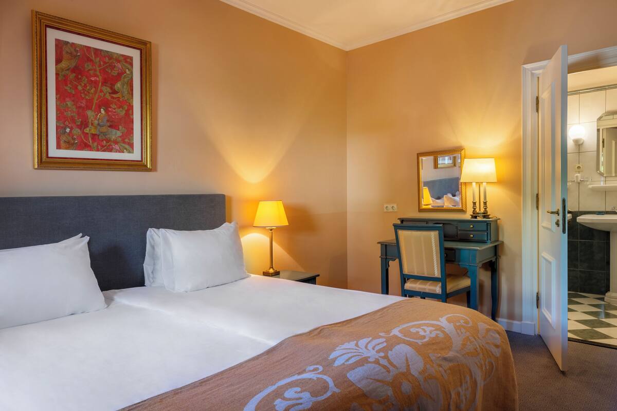 Une chambre d’hôtel avec un grand lit, un bureau avec une chaise, une lampe de bureau, un miroir et une salle de bain visible à travers une porte ouverte.