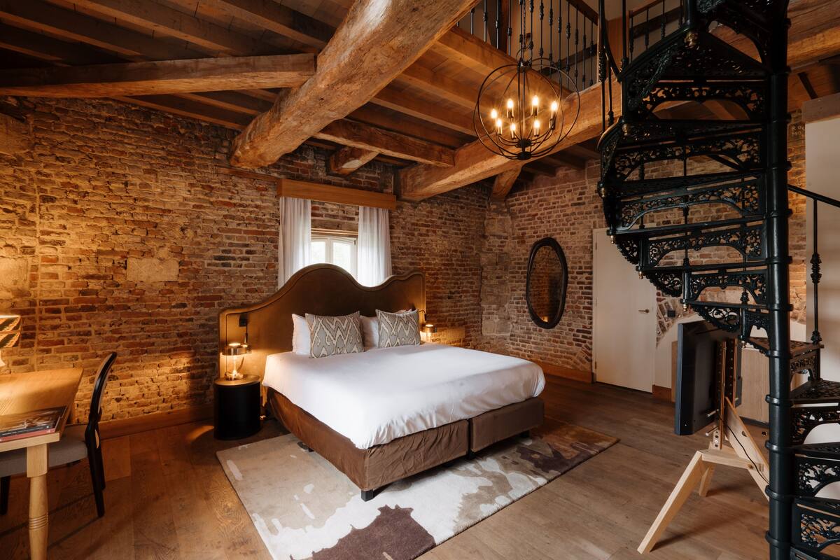 Une chambre à coucher rustique avec des murs en briques apparentes, un plafond en bois, un lit à baldaquin, un escalier en colimaçon et un bureau avec une lampe.