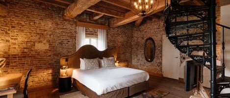 A rustic bedroom with exposed brick walls, a wooden ceiling, a four-poster bed, a spiral staircase, and a desk with a lamp.