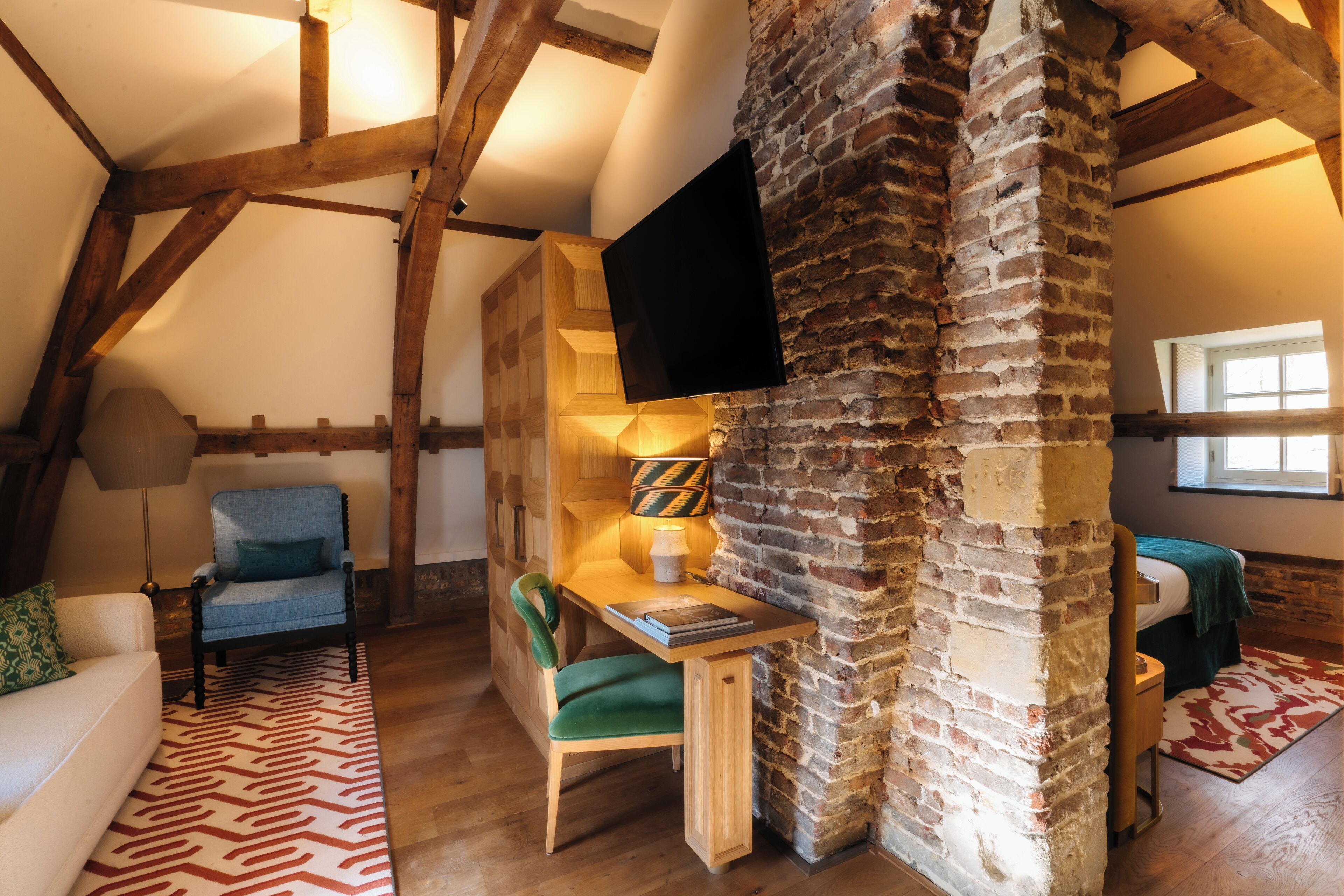 Ein gemütliches Dachzimmer mit freiliegenden Holzbalken, einer Ziegelwand, einem Flachbildfernseher, einem Schreibtisch mit Stuhl und einem Bett mit grüner Decke.