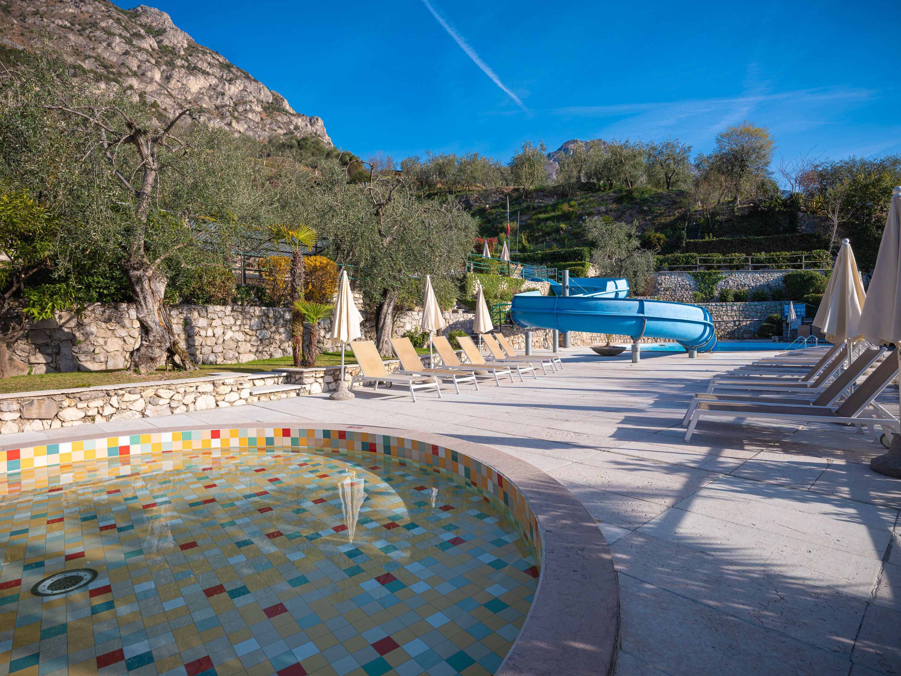 Un espace piscine agrémenté de chaises longues et de parasols, entouré de murs en pierre et d’arbres.