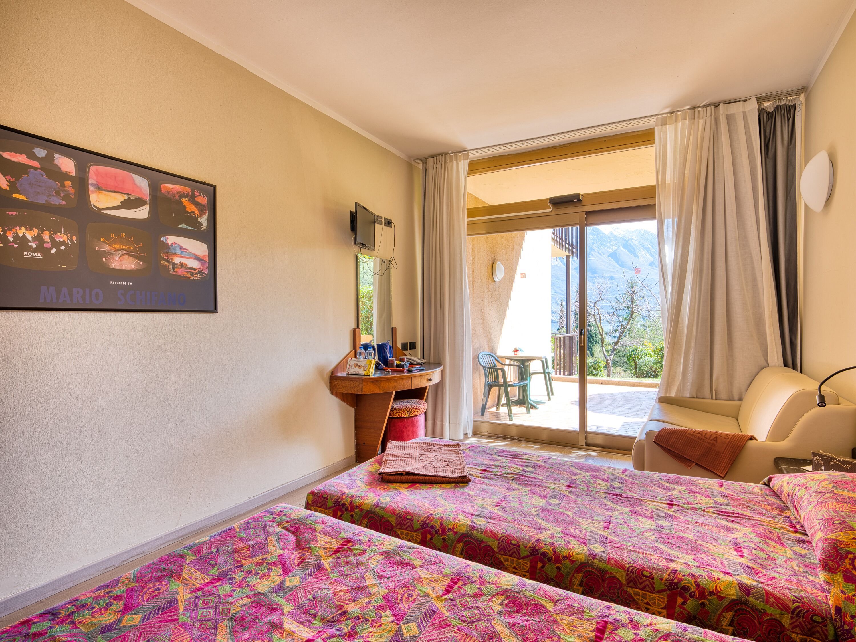 A hotel room with two beds, a desk with a chair, a window with curtains, and a view of mountains.