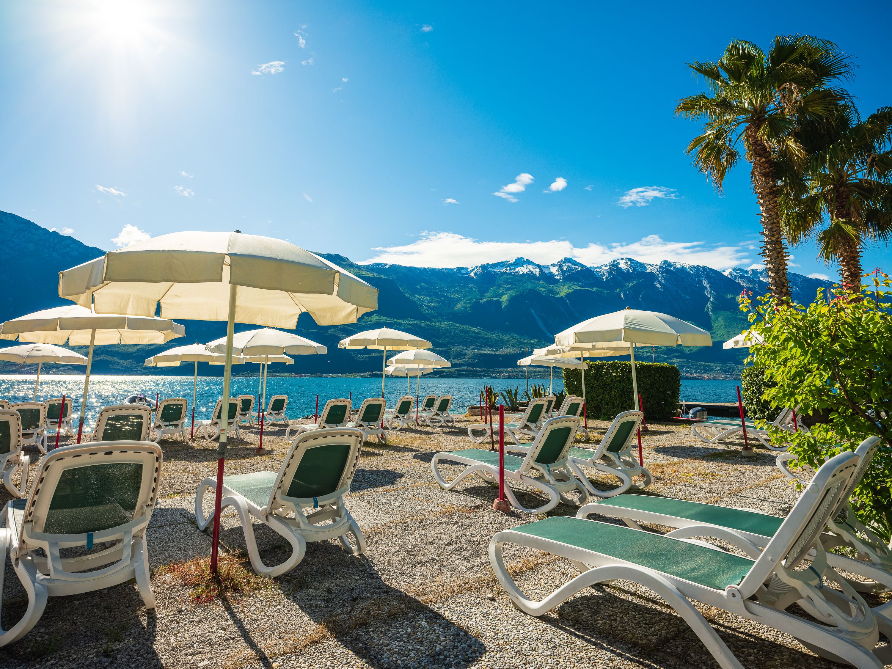 A beachside lounge with white lounge chairs and umbrellas, a pebble beach, and a lake with mountains in the background.