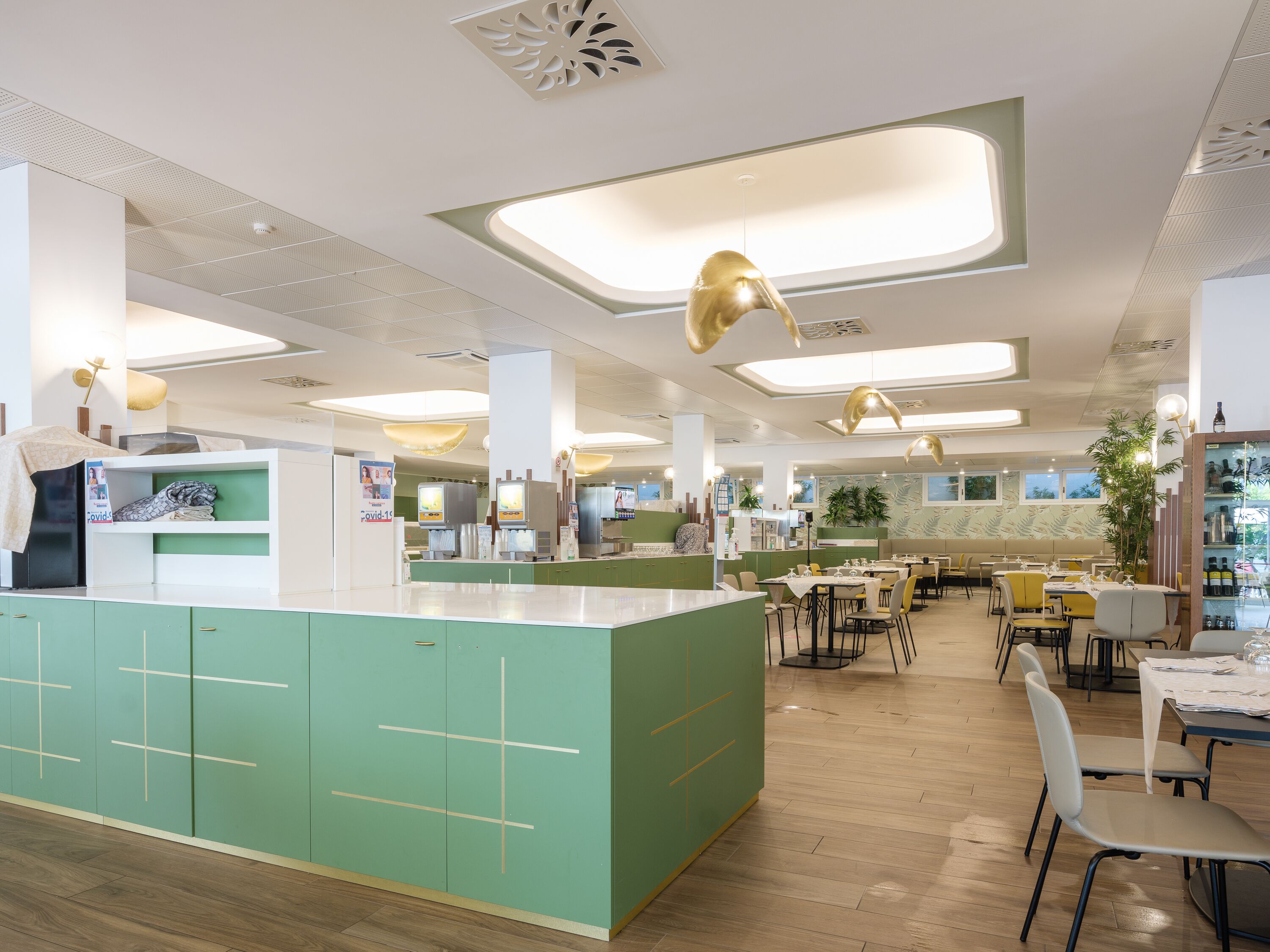 A modern restaurant interior with a central counter, green cabinetry, and multiple dining tables.