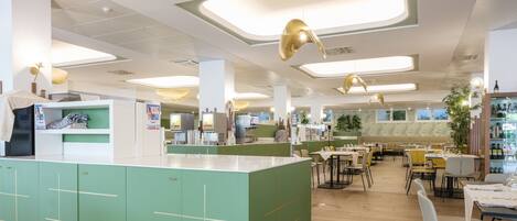 A modern restaurant interior with a central counter, green cabinetry, and multiple dining tables.