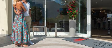 Entrada de un hotel con puerta de cristal, una planta grande en maceta con flores rojas y un felpudo con el nombre del hotel.