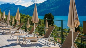 Una terraza con tumbonas y sombrillas, con vistas a un lago y montañas al fondo.