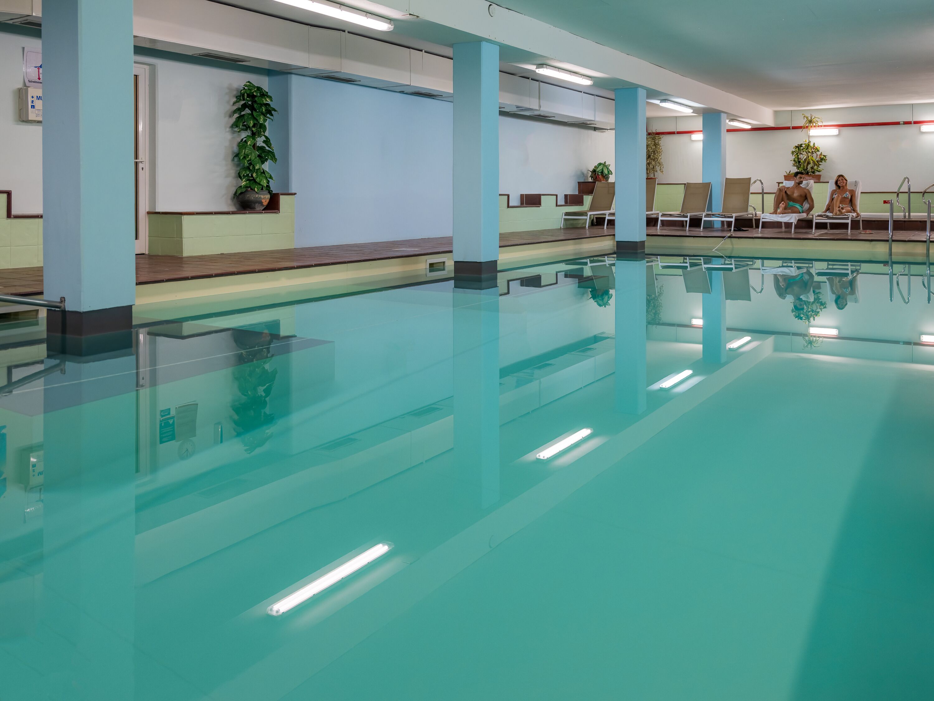 An indoor swimming pool with clear water, surrounded by light blue walls and columns, and equipped with overhead lighting.