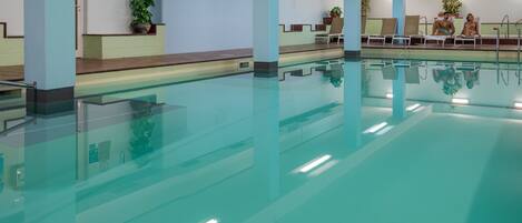 An indoor swimming pool with clear water, surrounded by light blue walls and columns, and equipped with overhead lighting.