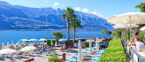 A seaside terrace with lounge chairs, umbrellas, and tables overlooking a body of water and mountains.