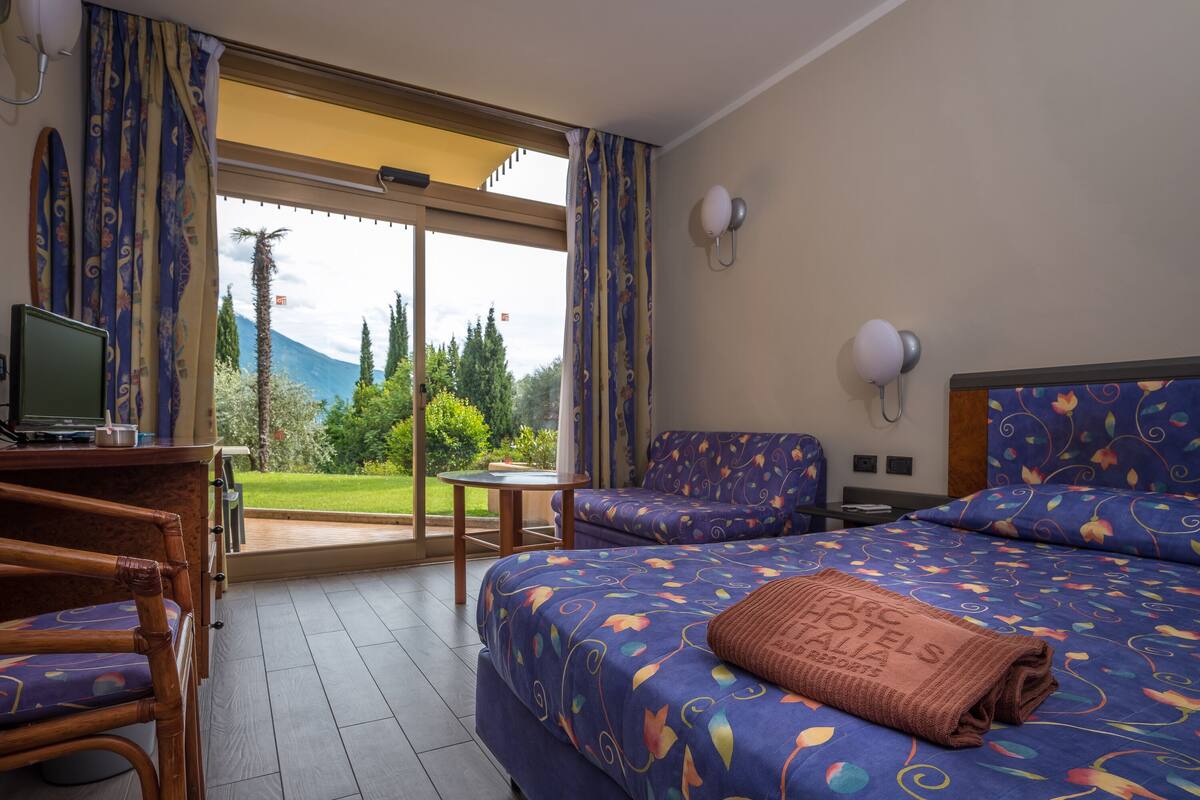 A hotel room with a bed, a desk, a chair, and a view of a garden and mountains.