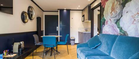 Una sala de estar moderna con un sofá azul, un comedor con sillas azules, un televisor montado en la pared y un cuadro grande con motivos florales.