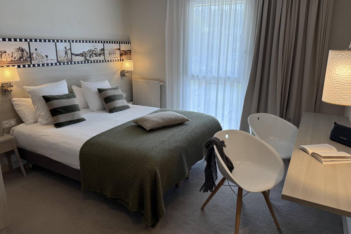 Un cuarto de hotel ordenado con cama, una silla, un escritorio y una lámpara.
