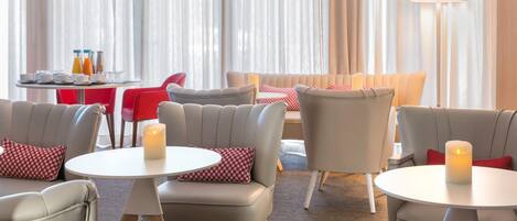A hotel lobby with round tables, grey and red chairs, and a large window with sheer curtains.