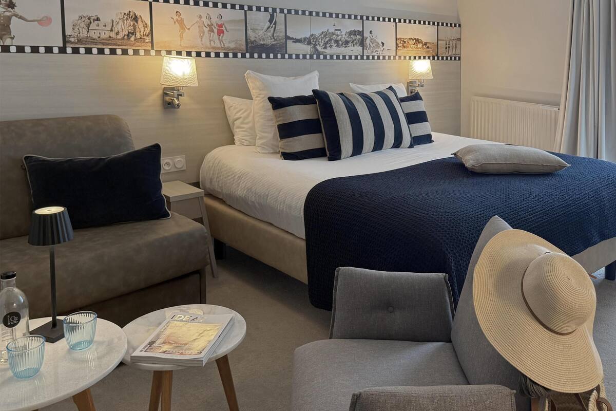 Una habitación de hotel con una cama, un sofá, una mesita con vasos y una revista, una lámpara y una pared decorada con una franja de fotografías en blanco y negro.