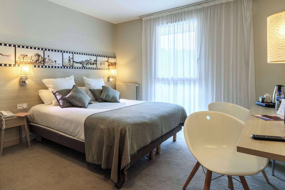 Una habitación de hotel con cama, mesitas de noche, un escritorio con silla, lámpara y un cuadro colgado en la pared.