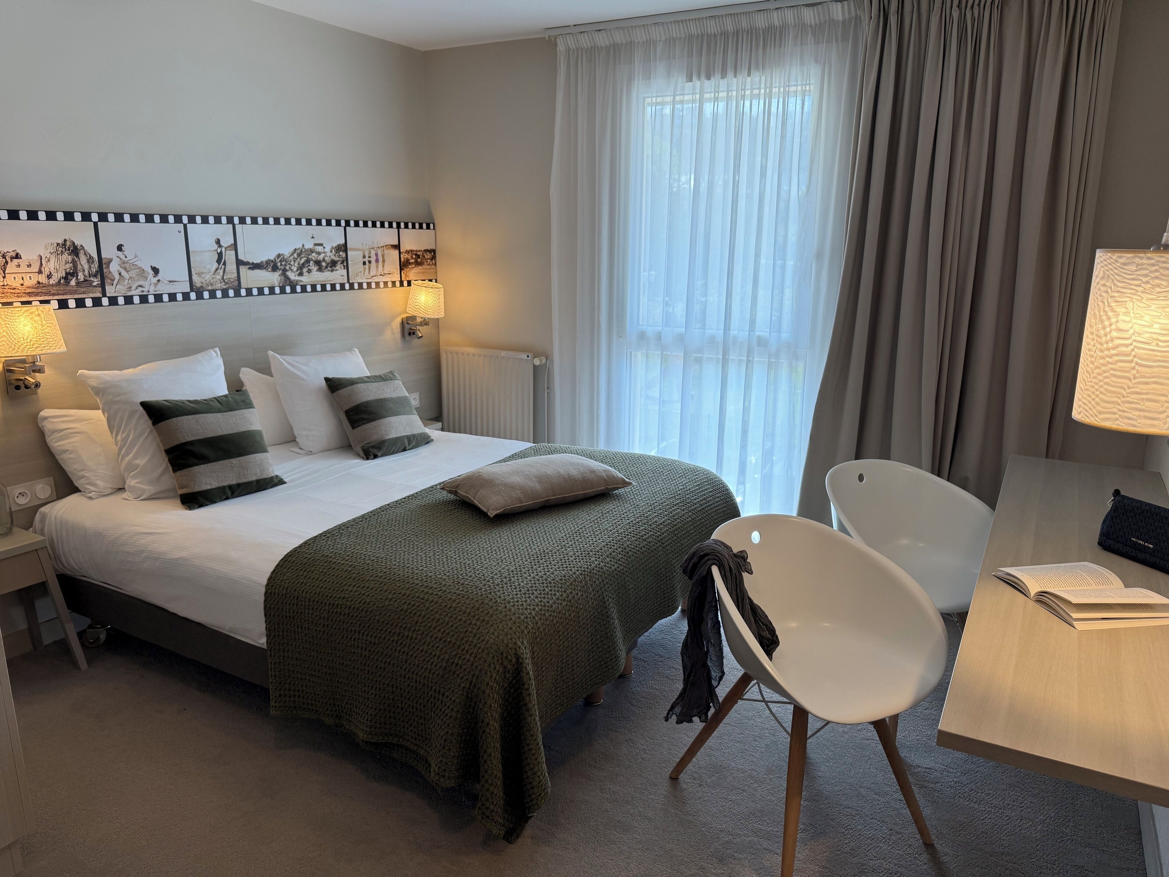 Une chambre d’hôtel avec un lit soigneusement fait, une chaise, un bureau avec une lampe et une vue sur l’extérieur à travers des rideaux légers.