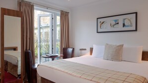 Ein Hotelzimmer mit einem großen Bett, einem Nachttisch, einem Sessel, einem Fenster mit Vorhängen und einem gerahmten Bild an der Wand.