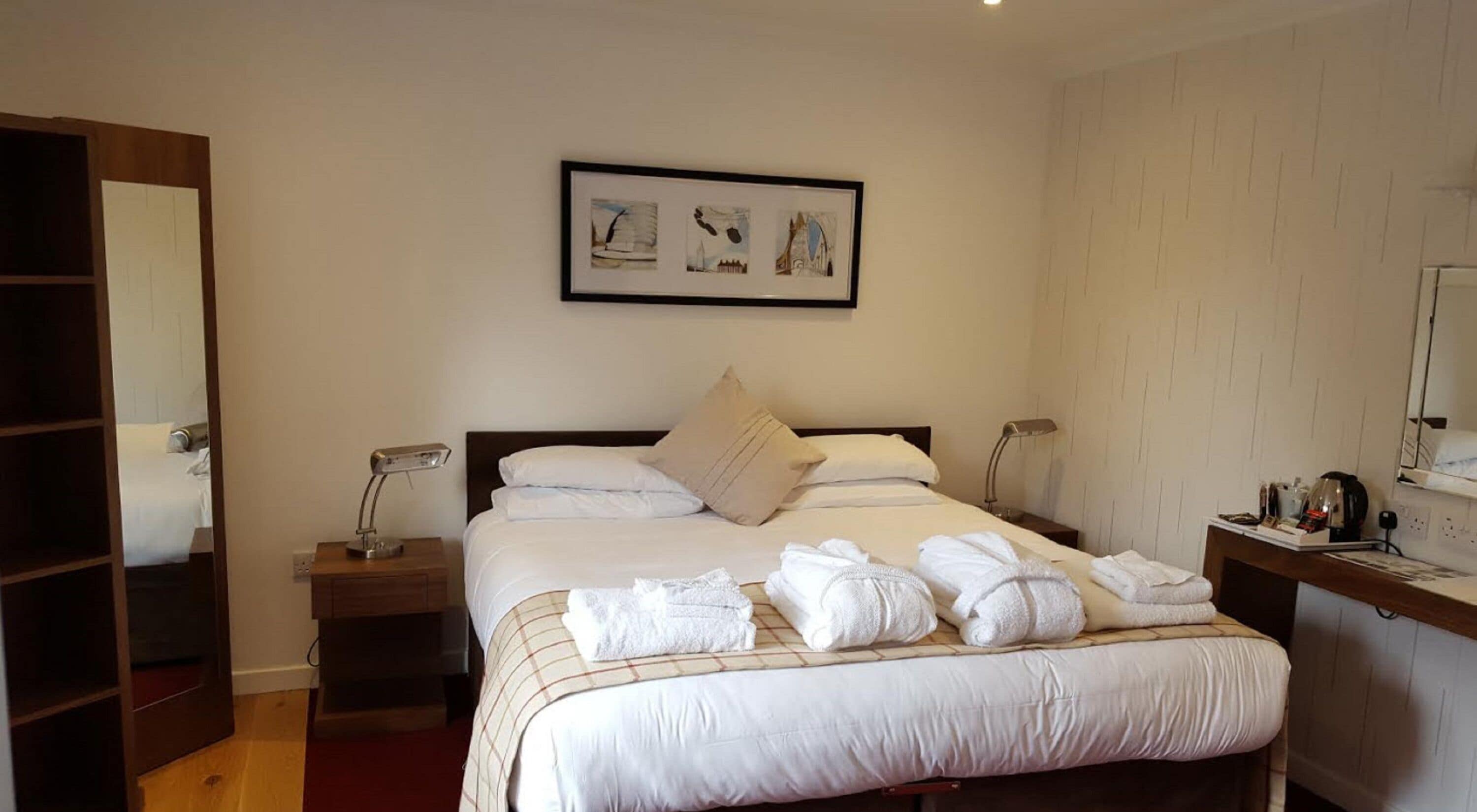 Habitación de hotel con una cama grande, una mesita de noche con lámpara, un espejo junto a la cama y un cuadro en la pared.