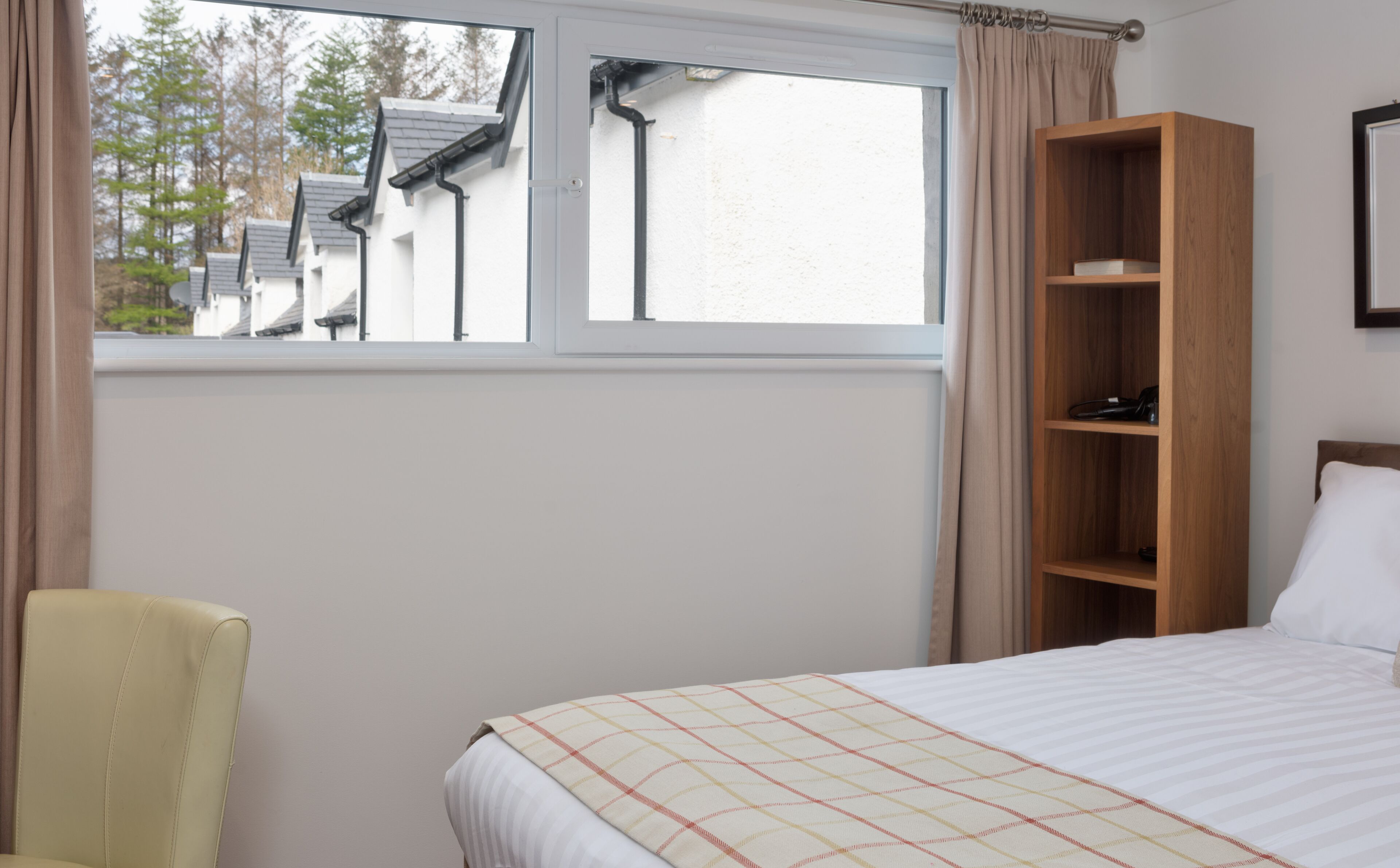 A bedroom with a bed, a wooden shelf, a chair, and a window with a view of houses and trees.